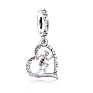 Könnte beinhalten: Ein silberner Charm-Anhänger mit herzförmigem Anhänger, der eine rennende Figur in Rot, Blau und Rosa zeigt. Das Herz ist mit "She believed she could so she did" beschriftet und mit klaren Kristallen verziert.