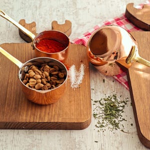 Puede incluir: Cacerolas de cobre con asas doradas, llenas de granos de café y especias rojas, sobre tablas de cortar de madera. Una gran cuchara de cobre descansa cerca. La escena se desarrolla sobre una superficie de madera clara.