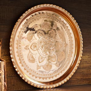 Puede incluir: Una bandeja redonda de color cobre con intrincados patrones florales y geom&eacute;tricos. La bandeja tiene un borde decorativo festoneado y se muestra sobre un fondo de madera oscura. El dise&ntilde;o presenta un motivo central en forma de estrella rodeado de adornos detallados.