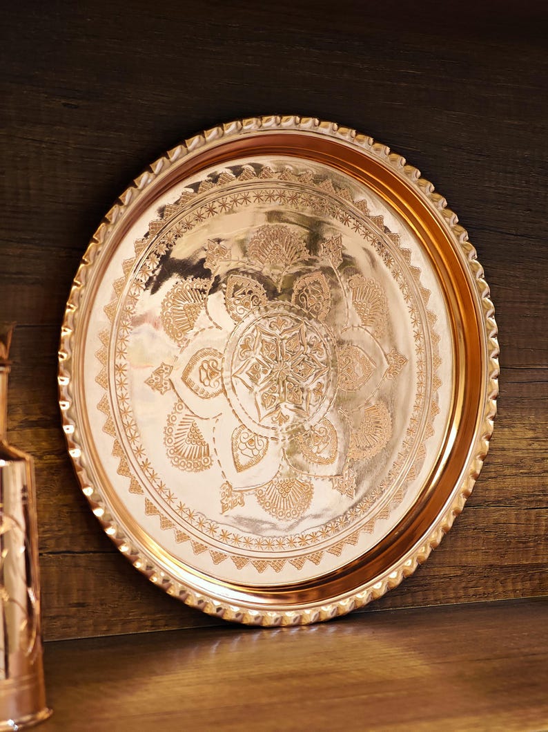 Puede incluir: Una bandeja redonda de color cobre con intrincados patrones florales y geom&eacute;tricos. La bandeja tiene un borde decorativo festoneado y se muestra sobre un fondo de madera oscura. El dise&ntilde;o presenta un motivo central en forma de estrella rodeado de adornos detallados.