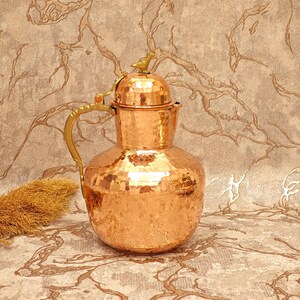 May include: A copper pitcher with a hammered finish, featuring a rounded base and a bird-shaped finial on the lid. It has a brass handle and spout. The copper has a reddish-brown colour, and the surface is textured with small hammer marks.