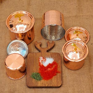 Puede incluir: Colección de utensilios de cocina de cobre, que incluye ollas pequeñas con tapas, un recipiente cilíndrico y una pequeña tabla de cortar con especias. Los artículos de cobre tienen una textura martillada y un acabado brillante.