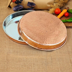 May include: A set of two round copper serving trays. The top tray is hammered and has a shiny finish. The bottom tray is smooth and has a reflective finish.