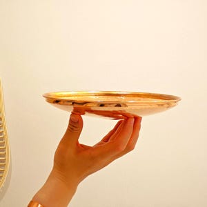 Könnte beinhalten: Eine kupferfarbene, flache Schale mit abgerundetem Rand. Die Schale wird von einer Hand gehalten. Die Schale hat eine reflektierende Oberfläche. Der Hintergrund ist eine schlichte, cremefarbene Wand. Links ist eine goldfarbene, fächerförmige Wanddekoration zu sehen.