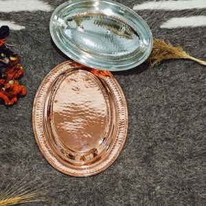 May include: Two oval serving trays. One is copper-colored with a hammered texture and a decorative rim. The other is clear glass. Both trays are displayed on a gray and white striped surface. The trays are empty.