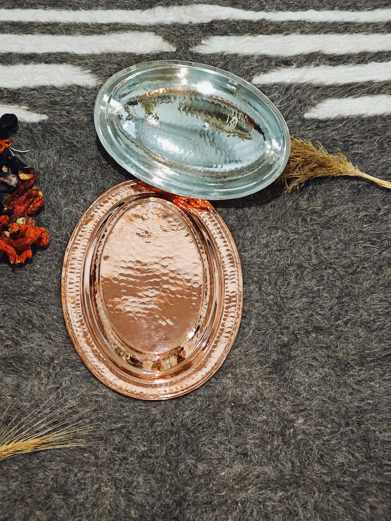 May include: Two oval serving trays. One is copper-colored with a hammered texture and a decorative rim. The other is clear glass. Both trays are displayed on a gray and white striped surface. The trays are empty.