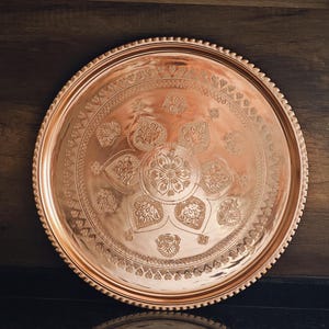 Puede incluir: Una bandeja redonda de cobre con intrincados patrones florales y geom&eacute;tricos grabados en su superficie. La bandeja tiene un borde con cuentas y un acabado pulido. Art&iacute;culos de cobre decorativos.
