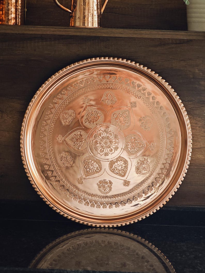 Puede incluir: Una bandeja redonda de cobre con intrincados patrones florales y geom&eacute;tricos grabados en su superficie. La bandeja tiene un borde con cuentas y un acabado pulido. Art&iacute;culos de cobre decorativos.
