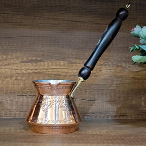 May include: A copper-colored Turkish coffee pot, also known as a cezve, with an ornate design. It has a long, black wooden handle and a brass-colored attachment. The pot is displayed on a wooden surface.