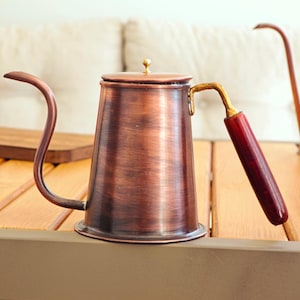 Puede incluir: Una tetera de cuello de cisne de color cobre con un mango de madera y un pequeño pomo de latón en la tapa. La tetera está sobre una mesa de madera. El pico se curva con gracia y el mango es de color rojo oscuro.