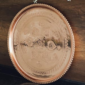 Puede incluir: Una bandeja redonda de cobre con un intrincado patr&oacute;n floral y geom&eacute;trico. La bandeja tiene un borde elevado con un borde de cuentas. El cobre tiene un acabado pulido. La bandeja se muestra sobre una superficie oscura.