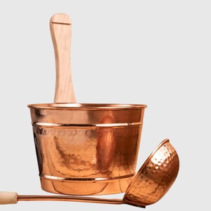 Puede incluir: Un cubo de sauna de cobre con un asa de madera y un cuchar&oacute;n a juego. El cubo tiene una textura martillada y el cuchar&oacute;n tiene un mango largo. Los art&iacute;culos est&aacute;n dise&ntilde;ados para su uso en una sauna.