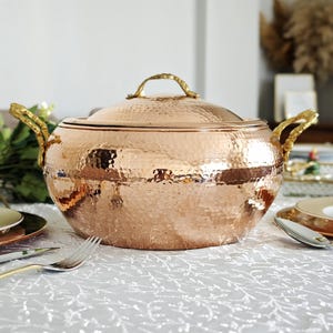 Peut inclure: Un grand plat de service en cuivre martelé avec un couvercle et deux poignées. Le plat est sur une nappe blanche à motif floral.