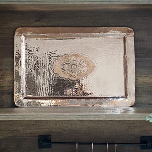 Könnte beinhalten: Ein rechteckiges Tablett aus Kupfer mit gehämmerten Texturen und einem dekorativen Blumendesign in der Mitte. Das Tablett, das auf einem Holzregal platziert ist, zeigt seinen metallischen Glanz und seine komplizierten Details. Ein dekoratives Stück.