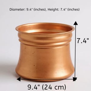 Może przedstawiać: Miedziany metalowy garnek o szerokim, zaokrąglonym korpusie i rozszerzonym brzegu. Garnek ma średnicę 24 cm i wysokość 19 cm. Garnek ma gładką, wypolerowaną powierzchnię i prosty, elegancki design.
