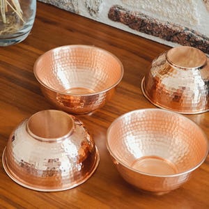 May include: Four copper bowls with a hammered texture are arranged on a wooden surface. The bowls have a warm, reddish-brown color and a reflective surface. Two bowls are open, and two are inverted.