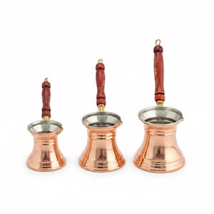May include: Three copper Turkish coffee pots with wooden handles. The pots have a shiny copper finish with a silver-colored interior and a wooden handle with a decorative pattern. The handles are attached with a brass-colored metal.