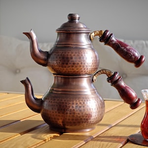 Puede incluir: Juego de té de cobre con una tetera de dos niveles y un vaso de té. La tetera tiene una textura martillada, una tapa y asas de madera. El té es de color rojo intenso y el juego está sobre una mesa de madera.