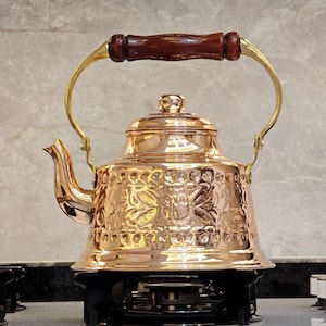 May include: A copper tea kettle with a decorative floral pattern. The kettle has a wooden handle and brass accents. The spout is curved, and the lid has a small knob. The kettle is sitting on a black stove top.