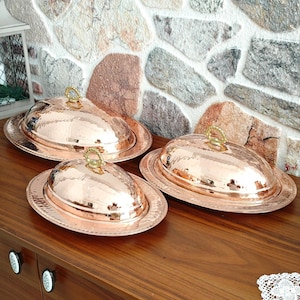 May include: Three copper serving dishes with lids. Each dish has an oval shape with a hammered texture and a gold-colored handle on the lid. The dishes are displayed on a wooden surface.