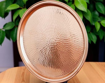 Puede incluir: Una bandeja redonda de metal color cobre con una superficie texturizada martillada. La bandeja tiene un borde decorativo con cuentas y se muestra sobre un soporte negro. El fondo presenta follaje verde y una superficie de madera.