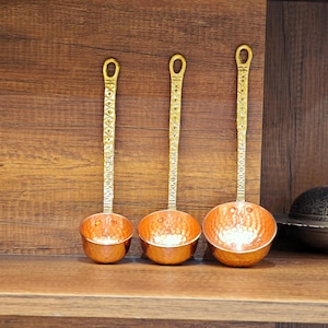 Puede incluir: Tres cucharones de cobre con mangos dorados y cuencos martillados descansan sobre una estantería de madera. Los mangos tienen un diseño ornamentado y los cuencos tienen un acabado brillante y reflectante. También se ve un cuenco decorativo de metal con tapa.