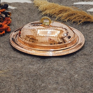 May include: A hammered copper serving dish with a lid. The dish has a round base and a domed lid with a decorative handle. The copper has a warm, reddish-brown color and a textured surface. The dish is designed for serving food.