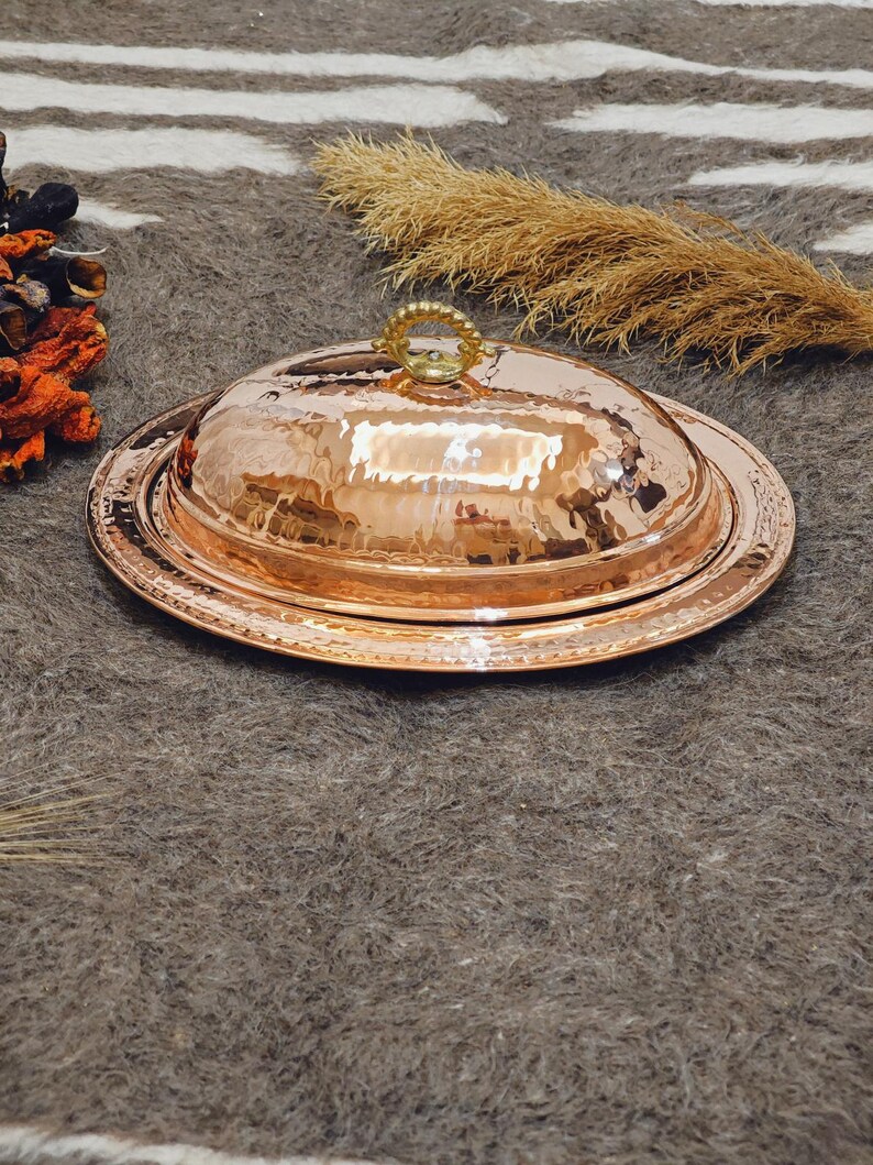 May include: A hammered copper serving dish with a lid. The dish has a round base and a domed lid with a decorative handle. The copper has a warm, reddish-brown color and a textured surface. The dish is designed for serving food.