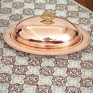 May include: A hammered copper serving dish with a domed lid and a decorative gold-colored handle. The dish sits on a white lace tablecloth, creating a contrast of textures and colors. The copper dish is oval-shaped.