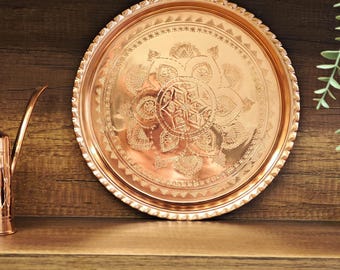 Puede incluir: Una bandeja redonda de cobre con intrincados diseños florales y geométricos. La bandeja tiene un borde decorativo y se exhibe en un estante de madera. La bandeja tiene aproximadamente 30 cm de diámetro.