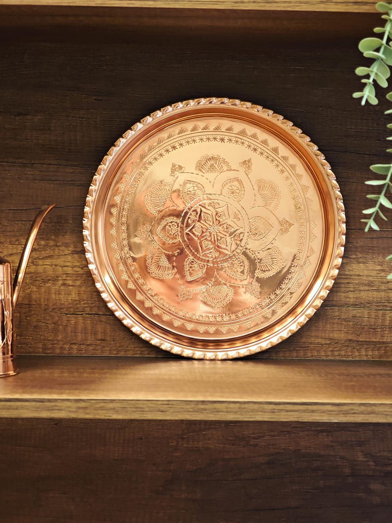 Puede incluir: Una bandeja redonda de cobre con intrincados dise&ntilde;os florales y geom&eacute;tricos. La bandeja tiene un borde decorativo y se exhibe en un estante de madera. La bandeja tiene aproximadamente 30 cm de di&aacute;metro.