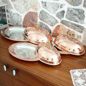 May include: Copper-colored serving dishes with lids. The lids have a hammered texture and a decorative handle. The serving platters have a silver interior and a copper-colored rim. The dishes are displayed on a wooden surface.