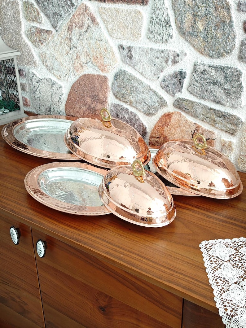 May include: Copper-colored serving dishes with lids. The lids have a hammered texture and a decorative handle. The serving platters have a silver interior and a copper-colored rim. The dishes are displayed on a wooden surface.