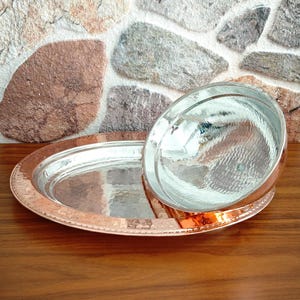 May include: A copper-colored serving tray with a hammered texture and a matching bowl. The oval tray has a raised rim and sits on a wooden surface. The bowl is round with a hammered texture and a shiny, reflective surface.