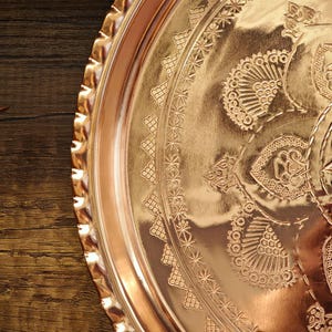 Puede incluir: Una bandeja redonda de color cobre con intrincados patrones florales y geom&eacute;tricos en relieve. La bandeja tiene un borde festoneado y un acabado pulido, que refleja la luz. El dise&ntilde;o incluye motivos detallados de estrellas y flores.