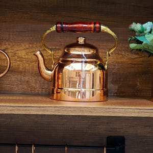 May include: A polished copper tea kettle with a wooden handle and a curved spout. The kettle is displayed on a wooden shelf. The handle is attached to the kettle with gold-coloured metal. A classic kitchenware item.