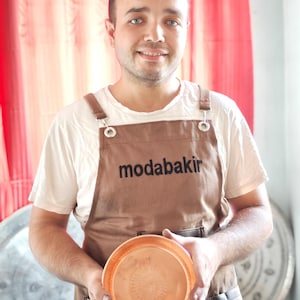 以下が含まれることがあります： 黒い文字で「modabakir」と書かれた茶色のエプロンを着た男性が、銅製のボウルを持っています。
