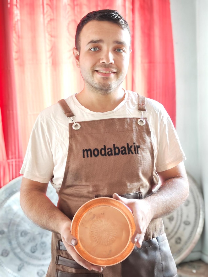 以下が含まれることがあります： 黒い文字で「modabakir」と書かれた茶色のエプロンを着た男性が、銅製のボウルを持っています。