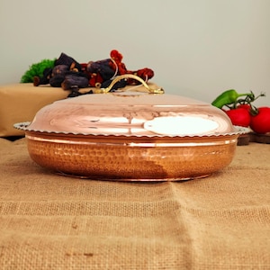 Può includere: Un piatto da portata in rame martellato con coperchio e manico dorato. Il piatto è appoggiato su una superficie di juta.