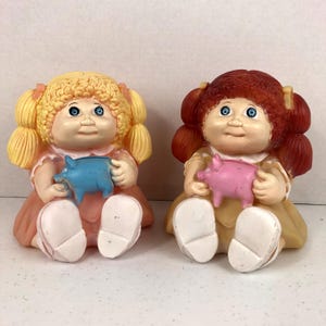 May include: Two Cabbage Patch Kid piggy banks. One is blonde with a pink dress and a blue piggy bank. The other is red-haired with a yellow dress and a pink piggy bank.