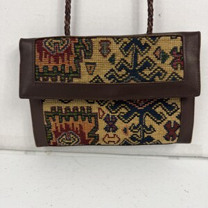 May include: A rectangular shoulder bag featuring a woven tapestry panel with geometric patterns in red, blue, green, and tan. The bag has a dark brown leather flap, matching leather trim, and a thin, braided dark brown leather shoulder strap.