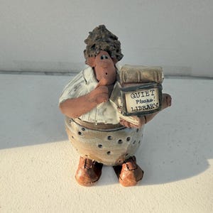 May include: A ceramic figurine of a person with dark curly hair, wearing a white shirt and a tan polka-dot skirt. The figure holds a book with a sign that reads QUIET Please! LIBRARY. The character has brown shoes and a finger raised to their mouth.