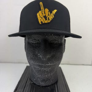 May include: A black baseball cap featuring a yellow embroidered patch on the front. The patch depicts a hand gesture with the middle finger raised, accompanied by the text ALOHA and the smaller text TWO JOINTS integrated into the design.