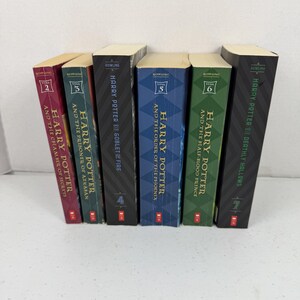 May include: A collection of six Harry Potter books standing upright in a row. The spines are colored maroon, teal, black, blue, green, and black, featuring the titles, author name Rowling, and year numbers two through seven in gold text.
