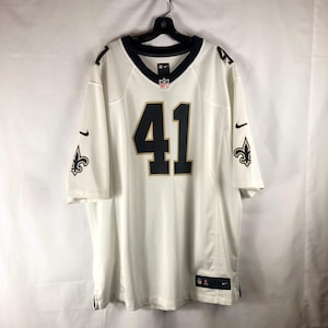 May include: White football jersey with black and gold accents. The jersey features the number 41 in black with a gold outline on the front. The sleeves have the Nike swoosh and a fleur-de-lis logo. The collar is black.