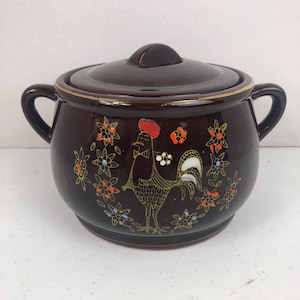 May include: A brown ceramic lidded pot with two handles. The pot is decorated with a rooster and floral design in gold, red, white, and blue. The lid has a small knob.