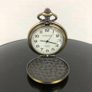 May include: An open, antique-style pocket watch with a white face and black numerals. The watch has a gold-colored metal casing and the word "STRADA" is printed on the face. The watch reads "QUARTZ JAPAN".