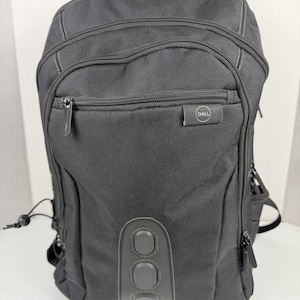 May include: A black Dell backpack featuring multiple zippered compartments, a front pocket with a small Dell logo tag, and a vertical grey accent panel at the base with three circular textured pads. The bag includes side straps and black zippers.
