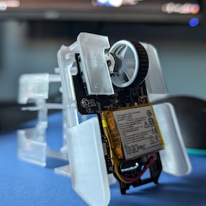 以下が含まれることがあります： 白のプラスチック製の3Dプリントケースで、内部に小さな回路基板が入っています。回路基板にはバッテリー、小型モーター、白黒の車輪が付いています。ケースは回路基板を保持し、保護するために設計されています。