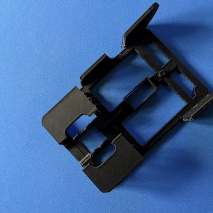 May include: A black plastic 3D printed frame with multiple slots. The frame is designed to hold small objects, such as SD cards or SIM cards.